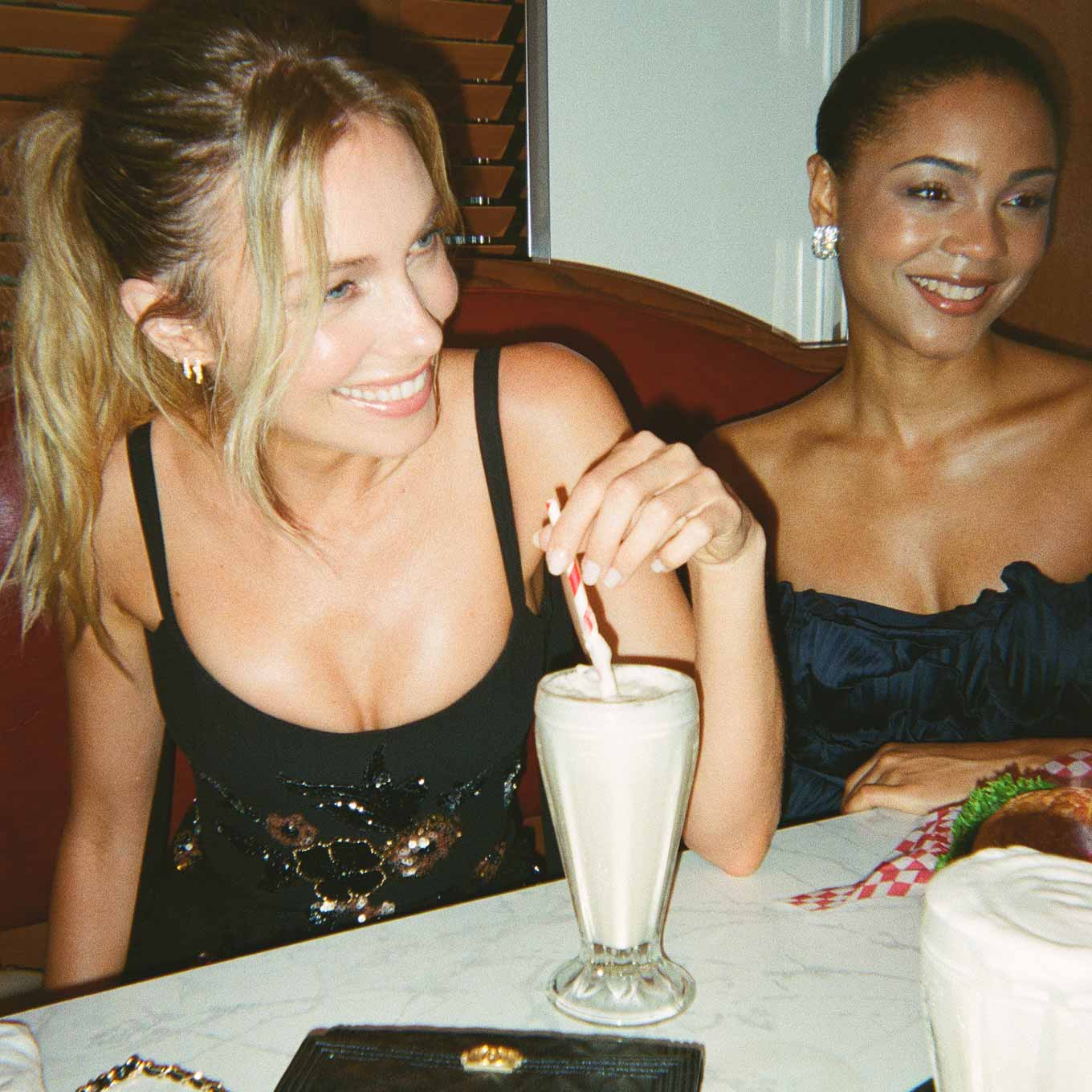 two women in eveningwear smiling at a table in a diner while one sips a milkshake