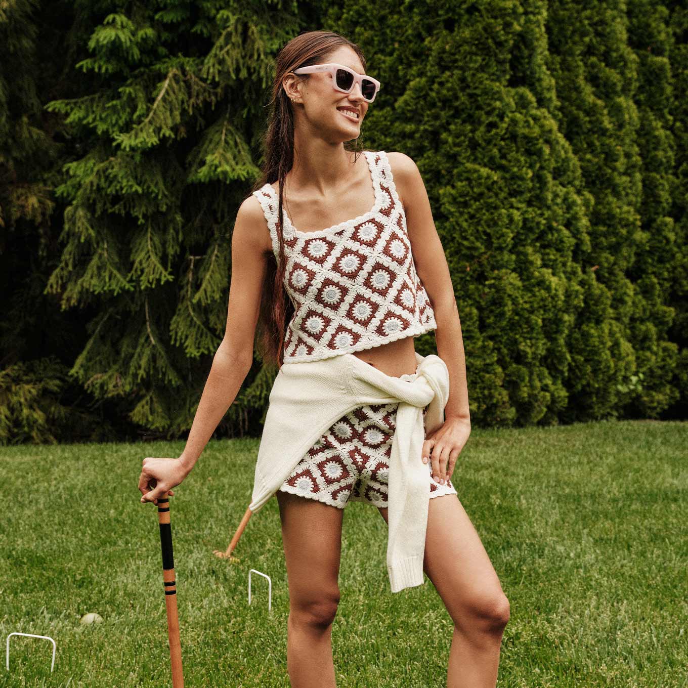 smiling woman wearing sunglasses and a crochet matching set in front of a croquet set